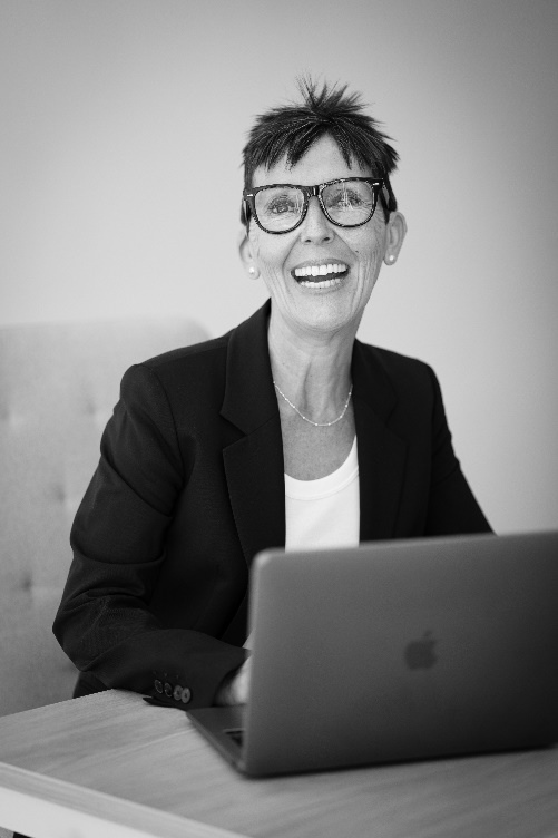 En blanco y negro vemos a una mujer de mediana edad sonriendo ampliamente a la cámara con un portátil abierto frente a ella. Lleva gafas de pasta, el pelo cortado de forma informal, mientras viste una camiseta y una chaqueta de traje