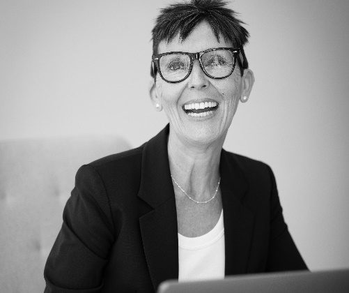 En blanco y negro vemos a una mujer de mediana edad sonriendo ampliamente a la cámara con un portátil abierto frente a ella. Lleva gafas de pasta, el pelo cortado de forma informal, mientras viste una camiseta y una chaqueta de traje