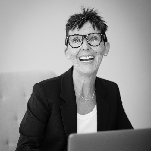 En blanco y negro vemos a una mujer de mediana edad sonriendo ampliamente a la cámara con un portátil abierto frente a ella. Lleva gafas de pasta, el pelo cortado de forma informal, mientras viste una camiseta y una chaqueta de traje